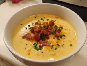 A close-up of a bowl of rich, yellow cheese soup topped generously with crumbled bacon bits and fresh green chives.