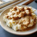 A close-up of tender chicken pieces smothered in creamy, seasoned chicken and gravy served over a mound of fluffy mashed potatoes.