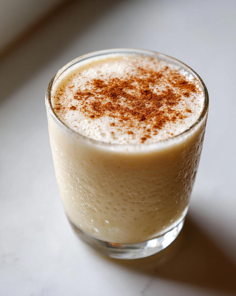 Close-up of a glass filled with creamy, chilled coquito, dusted generously with ground cinnamon on top.