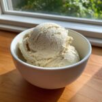 Two scoops of creamy, light-colored cottage cheese ice cream served in a white bowl on a wooden surface.