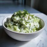 A white bowl filled with creamy dill pickle dip, featuring chunks of pickle and topped generously with fresh dill.