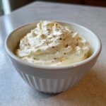 A close-up of a small white bowl filled with fluffy, creamy horseradish sauce, topped with cracked black pepper.