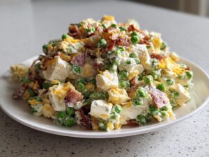 A generous serving of creamy pea salad mixed with chunks of cheese, bacon bits, and shredded cheddar on a white plate.