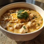 A close-up of a white bowl filled with creamy white chicken chili, topped with shredded cheese and fresh cilantro.