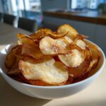 A white bowl overflowing with golden, crispy homemade potato chips sprinkled with coarse salt.