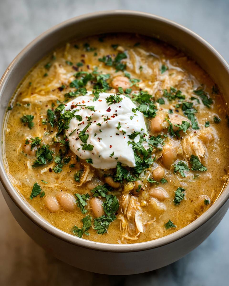 Close-up of a bowl of creamy crockpot white chicken chili topped with sour cream, cilantro, and chili flakes.