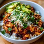 A close-up of a delicious chicken burrito bowl layered with rice, shredded chicken in sauce, black beans, cheese, avocado, and cilantro.