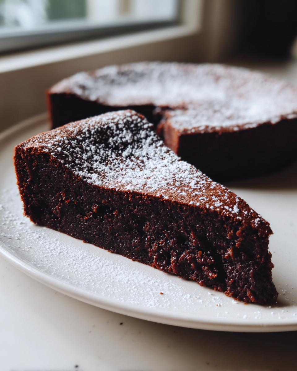 A rich, dark slice of flourless chocolate torte dusted with powdered sugar, sitting on a white plate.