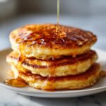 Close-up of a tall stack of golden brown banana pancakes being drizzled with syrup.