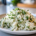 A close-up of fluffy cilantro lime rice piled high on a white plate, garnished generously with chopped green herbs.
