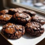 A pile of fudgy brownie cookies topped with flaky sea salt, sitting on a white plate.