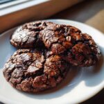 Three fudgy chocolate brownie cookies with cracked tops and visible chocolate chips resting on a white plate.