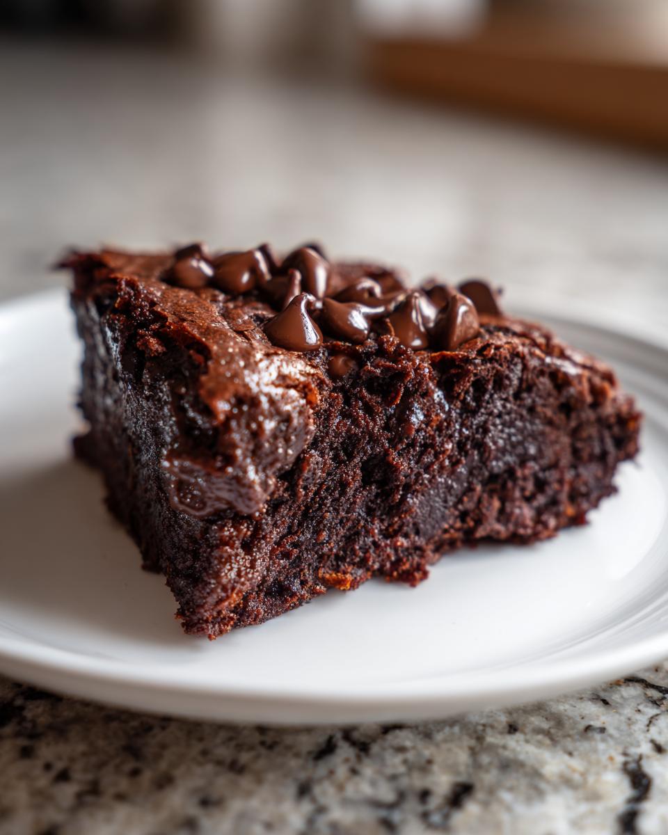 A close-up of a rich, dark chocolate slice of sweet potato brownies topped with melted chocolate chips on a white plate.