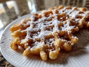 Close-up of a golden brown funnel cake recipe creation dusted heavily with powdered sugar on a white plate.