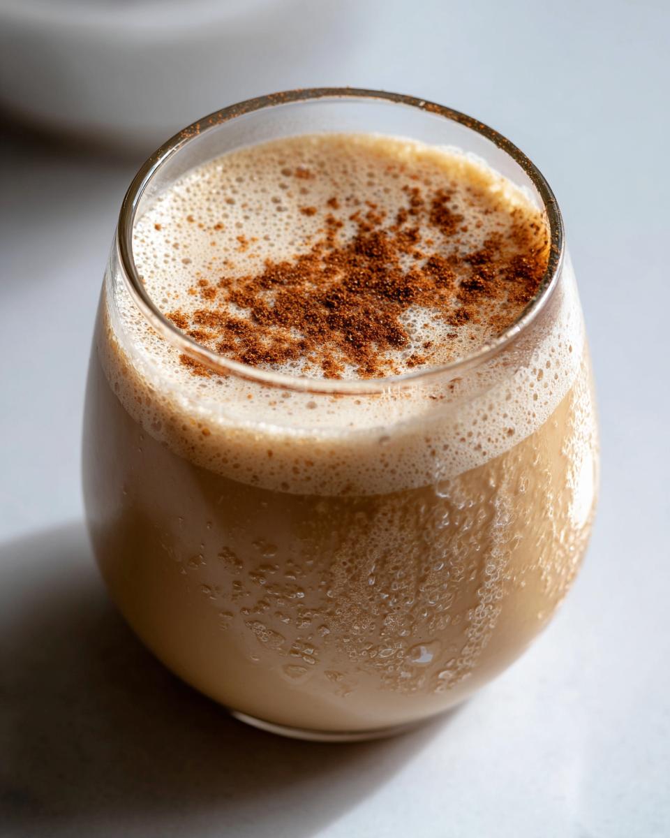 Close-up of a glass filled with creamy, chilled coquito topped with frothy foam and a dusting of ground cinnamon.