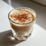 A small glass filled with creamy, frothy coquito, dusted generously with ground cinnamon.