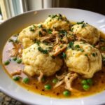 A close-up of a bowl of gluten free chicken and dumplings, featuring four large dumplings in savory broth with shredded chicken and peas.