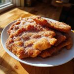 A stack of freshly fried, golden brown buñuelos generously coated in white sugar, resting on a white plate.