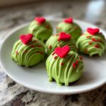 Close-up of six bright green Grinch Oreo Balls topped with red heart sprinkles on a white plate.