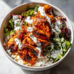 Close-up of a bowl featuring bright red harissa chicken pieces over white rice, mixed greens, and drizzled with a white yogurt sauce.