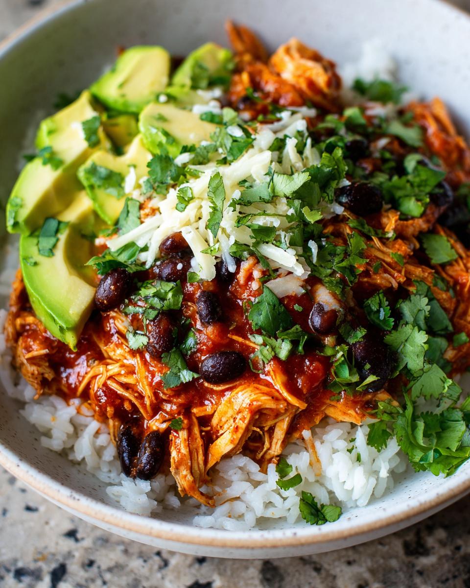 Close-up of a delicious chicken burrito bowl featuring shredded chicken in sauce, black beans, rice, avocado slices, cheese, and cilantro.