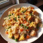 A close-up of homemade chicken fried rice mixed with scrambled egg, peas, and carrots, served on a white plate.