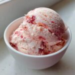 Two scoops of creamy, pink strawberry ice cream with visible fruit chunks served in a small white bowl.