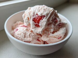 Close-up of several scoops of pale pink strawberry ice cream with visible chunks of red fruit in a white bowl.