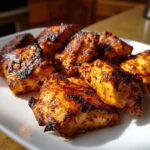 Close-up of perfectly grilled, charred chicken pieces coated in a rich, orange-red chicken marinade, served on a white plate.