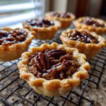 A close-up shot of several freshly baked mini pecan pies cooling on a wire rack, featuring gooey filling and whole pecans.