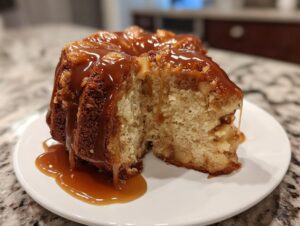A slice cut from a moist apple dapple cake, generously topped with dripping caramel sauce, served on a white plate.