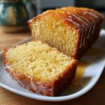 A close-up of a moist orange juice cake loaf with one slice cut, drizzled with a bright orange glaze.