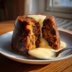 A rich, dark plum pudding cut open, generously topped with warm, flowing custard sauce.