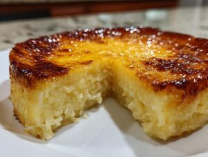 A close-up of a rich, golden-brown topped cassava cake sitting on a white plate with a piece removed.