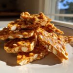 Close-up of broken pieces of homemade peanut brittle recipe stacked on a light-colored plate.