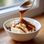 A spoonful of rich, thick homemade brown gravy is being poured over a mound of mashed potatoes in a white bowl.