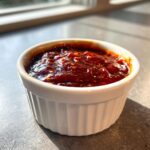 Close-up of thick, dark red homemade bbq sauce with visible spices in a small white ramekin.