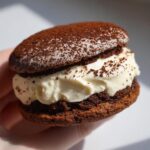 A close-up view of a delicious tiramisu cookies sandwich held in a hand, featuring dark chocolate cookies and creamy filling dusted with cocoa.