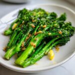 Close-up of bright green roasted broccolini tossed with crispy garlic and lemon zest on a white plate.