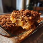 A spoonful of warm apple crisp, showing soft baked apples and crunchy oat topping, lifted from a glass baking dish.