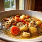 Close-up of a white bowl filled with rich, brothy slow cooker chicken stew featuring chunks of chicken, potatoes, and bright orange carrots.