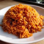 A close-up, appetizing mound of vibrant orange-red jollof rice served on a white plate.