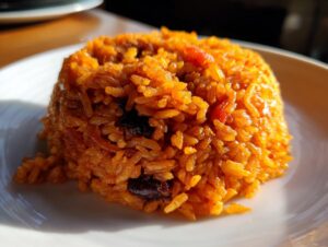 A close-up, brightly lit portion of vibrant orange jollof rice molded into a mound on a white plate.