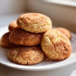 A close-up stack of freshly baked, soft chewy snickerdoodles coated in cinnamon sugar on a white plate.
