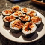 A plate of freshly cut spicy tuna kimbap rolls, showing the rice, seaweed, and vibrant orange tuna filling sprinkled with sesame seeds.