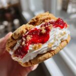 A hand holding one of the decadent strawberry cheesecake cookies, filled with cream and bright red strawberry topping.
