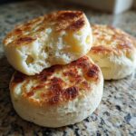 Three homemade english muffins stacked on a granite counter, showing the nooks and crannies texture of the toasted english muffin.