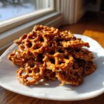 A mound of homemade toffee pretzels coated in a shiny caramel glaze, sitting on a white plate near a window.