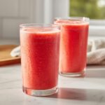 Two tall, sweating glasses filled with bright red, icy watermelon smoothie sitting on a white marble counter.