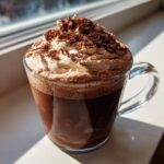 A clear glass mug filled with rich whipped hot chocolate, topped high with whipped cream and dusted generously with cocoa powder.